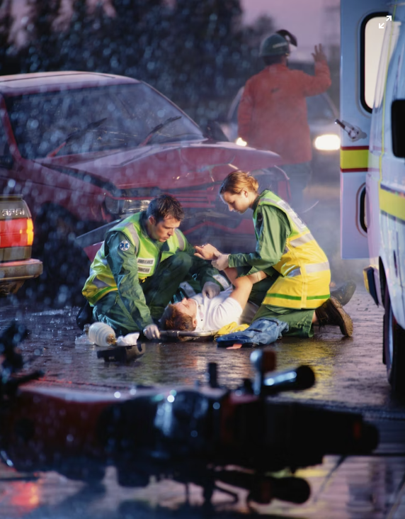 Paramedics aid an injured person on wet pavement by an ambulance after a road accident in the rain.