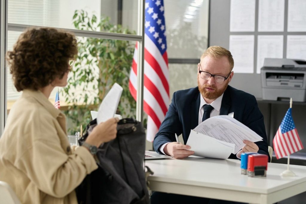 Man in suit reviews documents at a desk with a woman, American flags and office supplies visible.