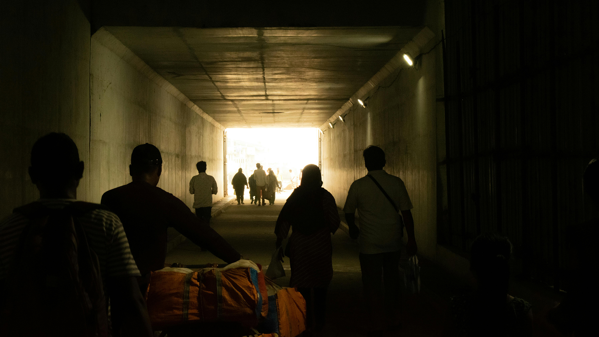 People walk through a dimly lit tunnel toward bright daylight at the tunnel’s exit.