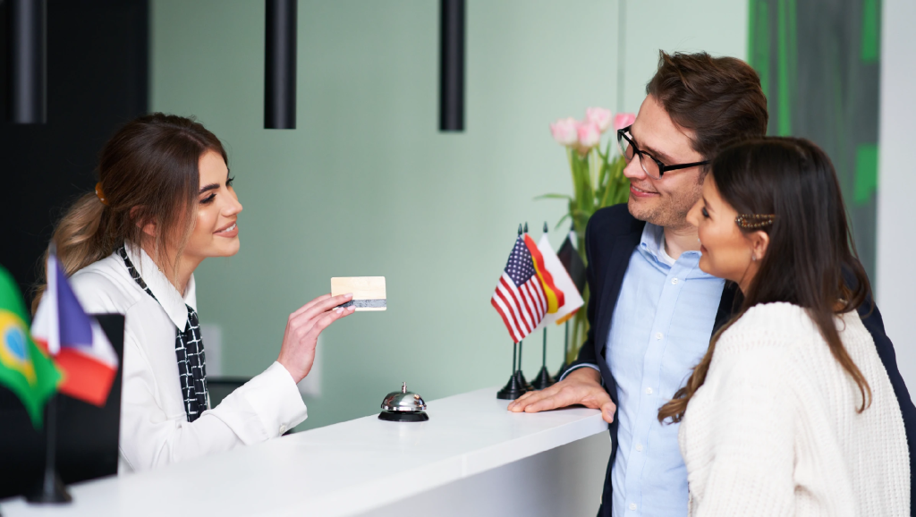 Hotel receptionist holding a key card for a smiling couple at check-in; small flags on the counter.