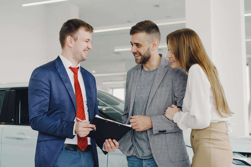 Car salesperson showing documents to a smiling couple in a showroom, standing in front of a car.
