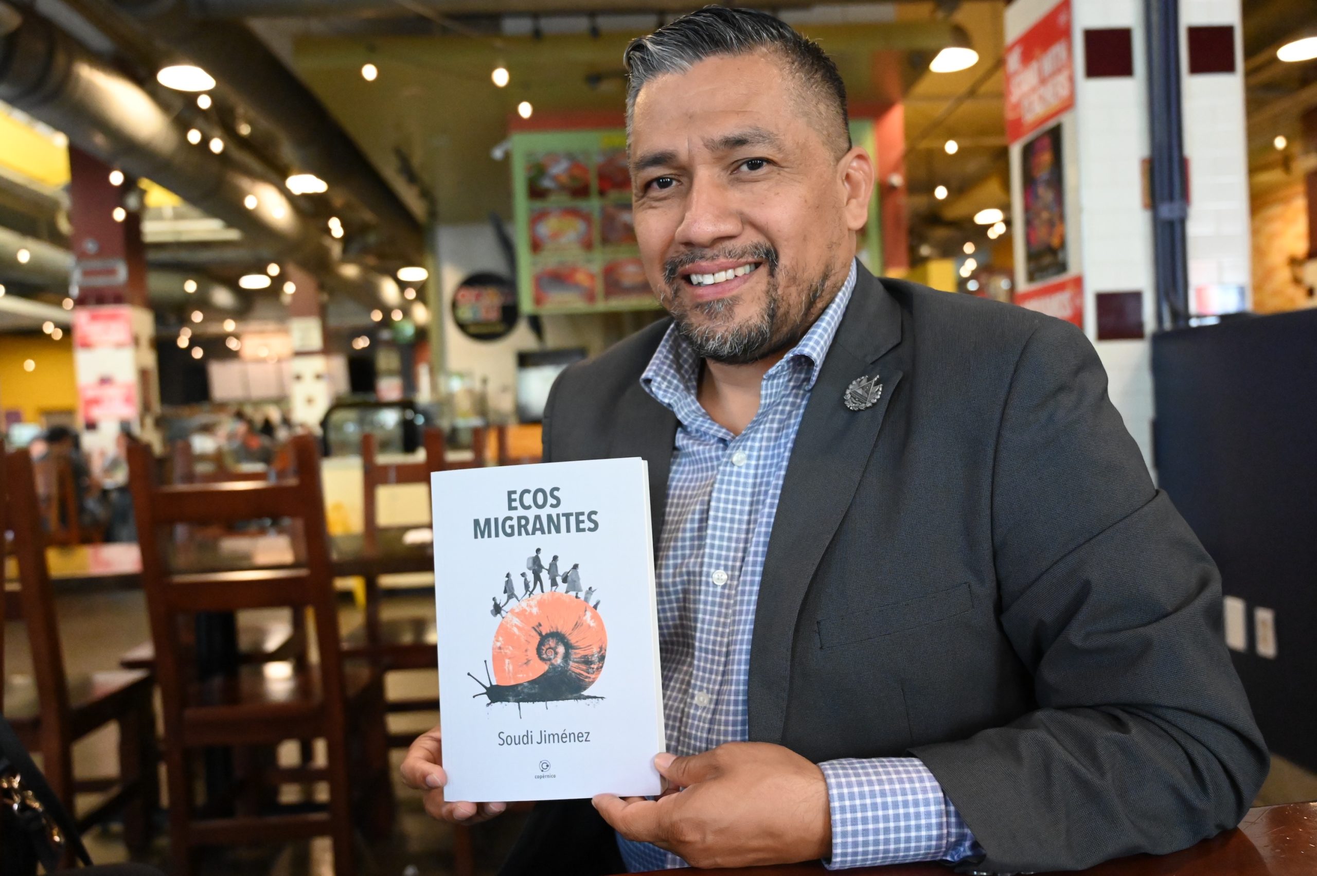 Smiling man in a suit holds up a book titled "Ecos Migrantes" by Soudi Jiménez in a restaurant setting, highlighting stories connected to Estados Unidos.