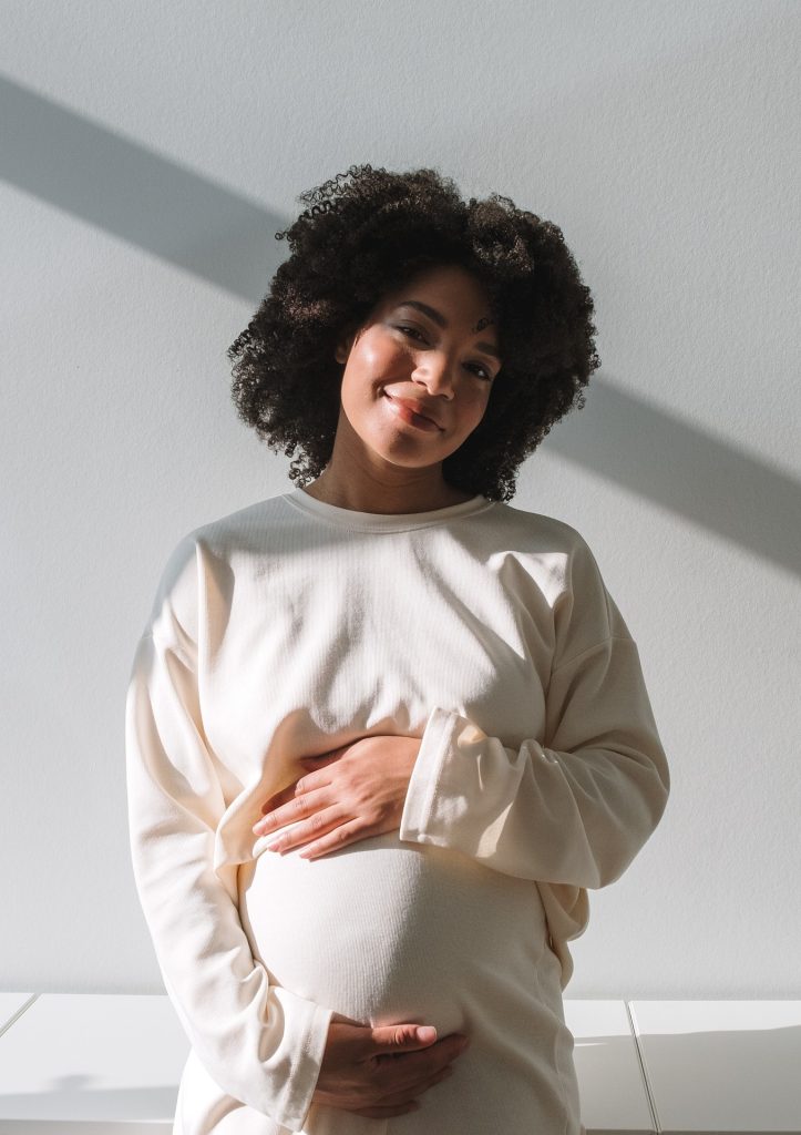 Pregnant woman in a cream sweater smiling and holding her belly while standing in sunlight against a white wall.