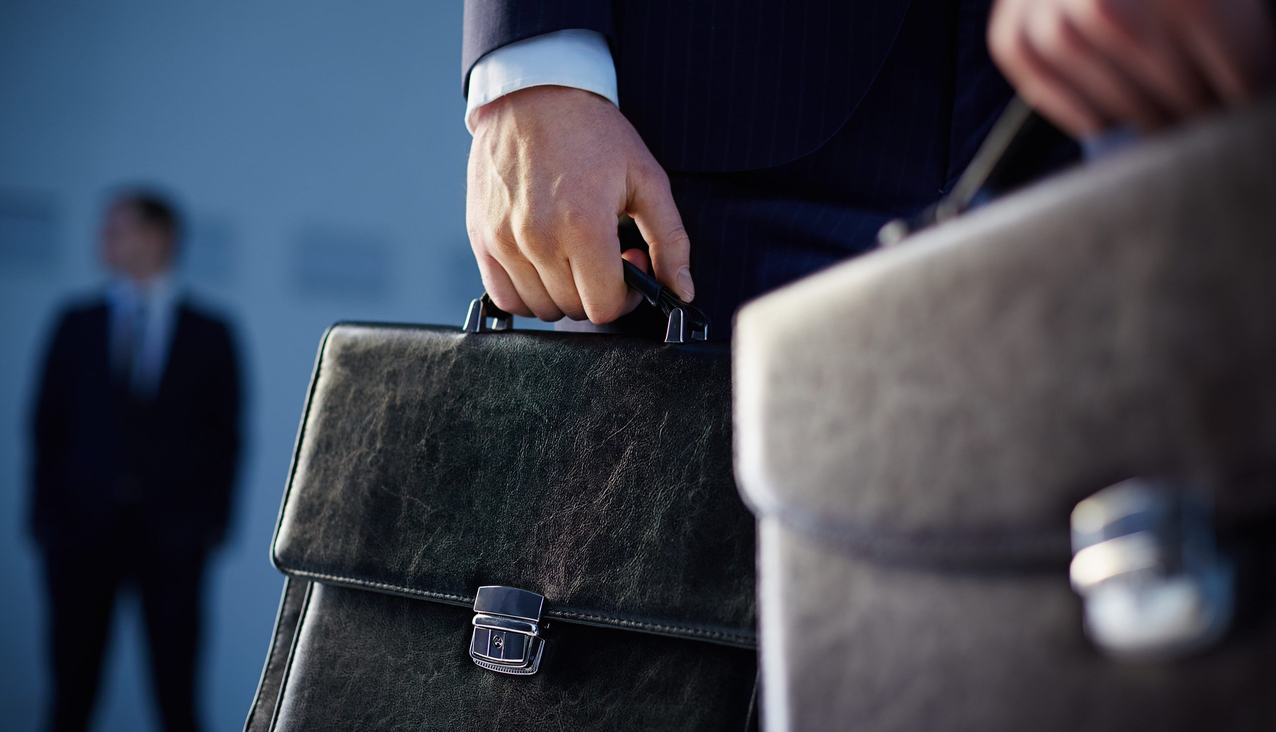 Close-up of two people in suits holding briefcases, with one person blurred in the background.
