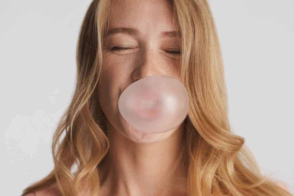 Smiling woman with long hair blowing a bubble with gum, eyes closed, against a plain background.