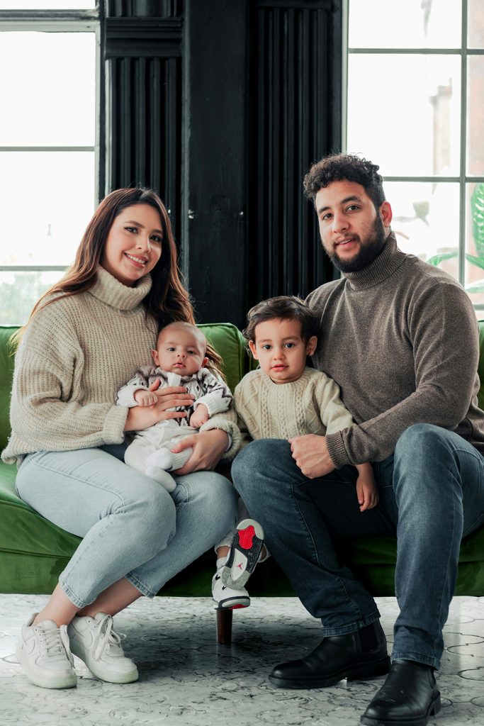 A family of four sits on a green couch, smiling in front of large windows.