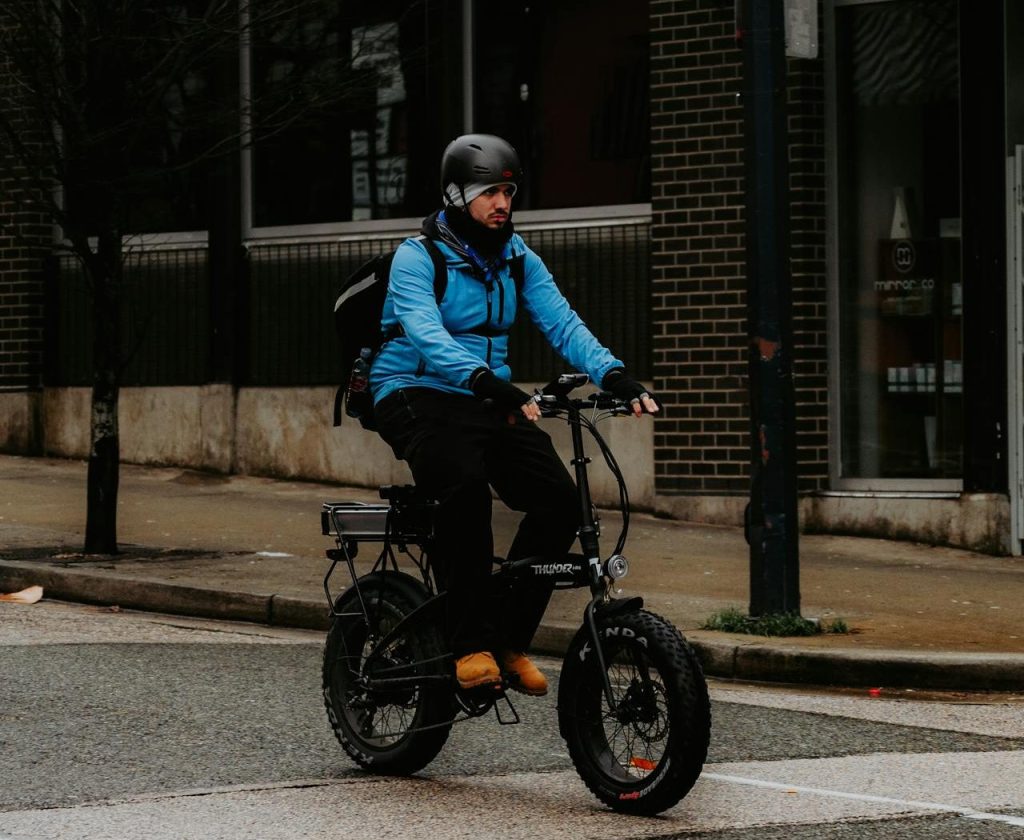 Person in a blue jacket and helmet riding an electric bike on a city street near a building and arrow sign.
