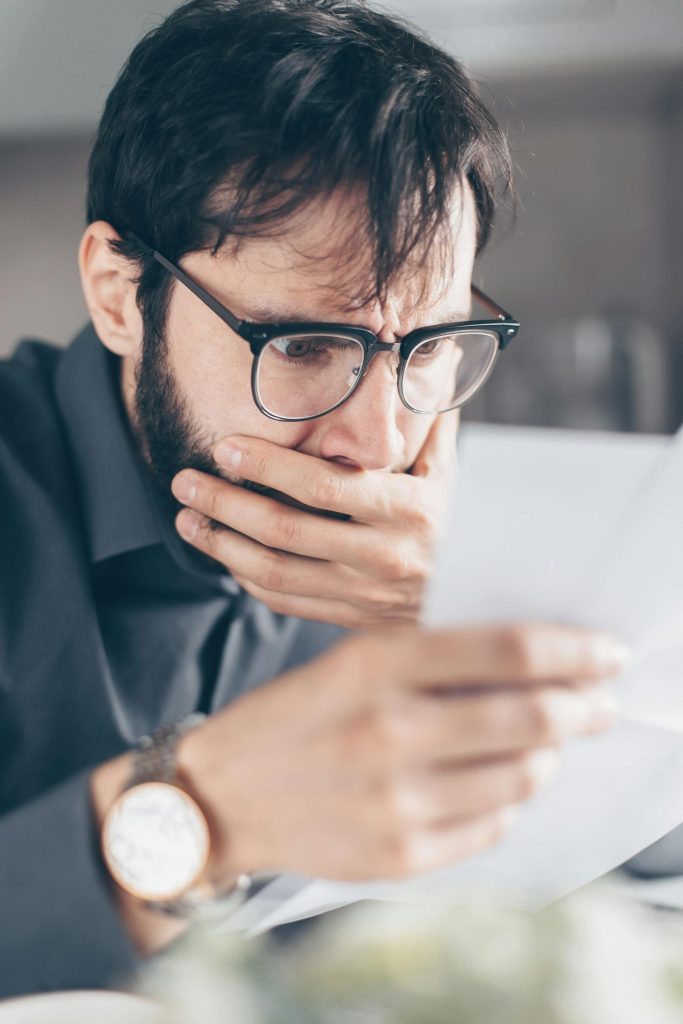 A man with glasses looks worried while reading a piece of paper, covering his mouth with his hand.
