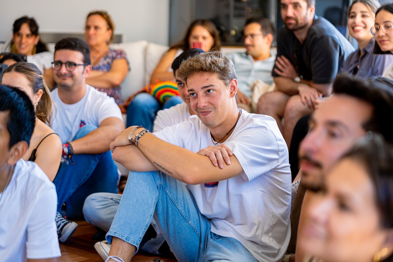 A group of people sit closely together on the floor, smiling and listening attentively.