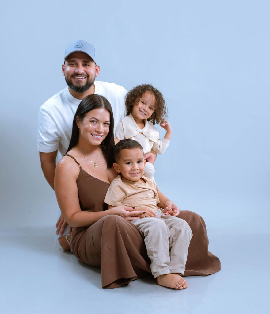 A smiling family of four poses together on a light blue background, with two children in front of their parents.
