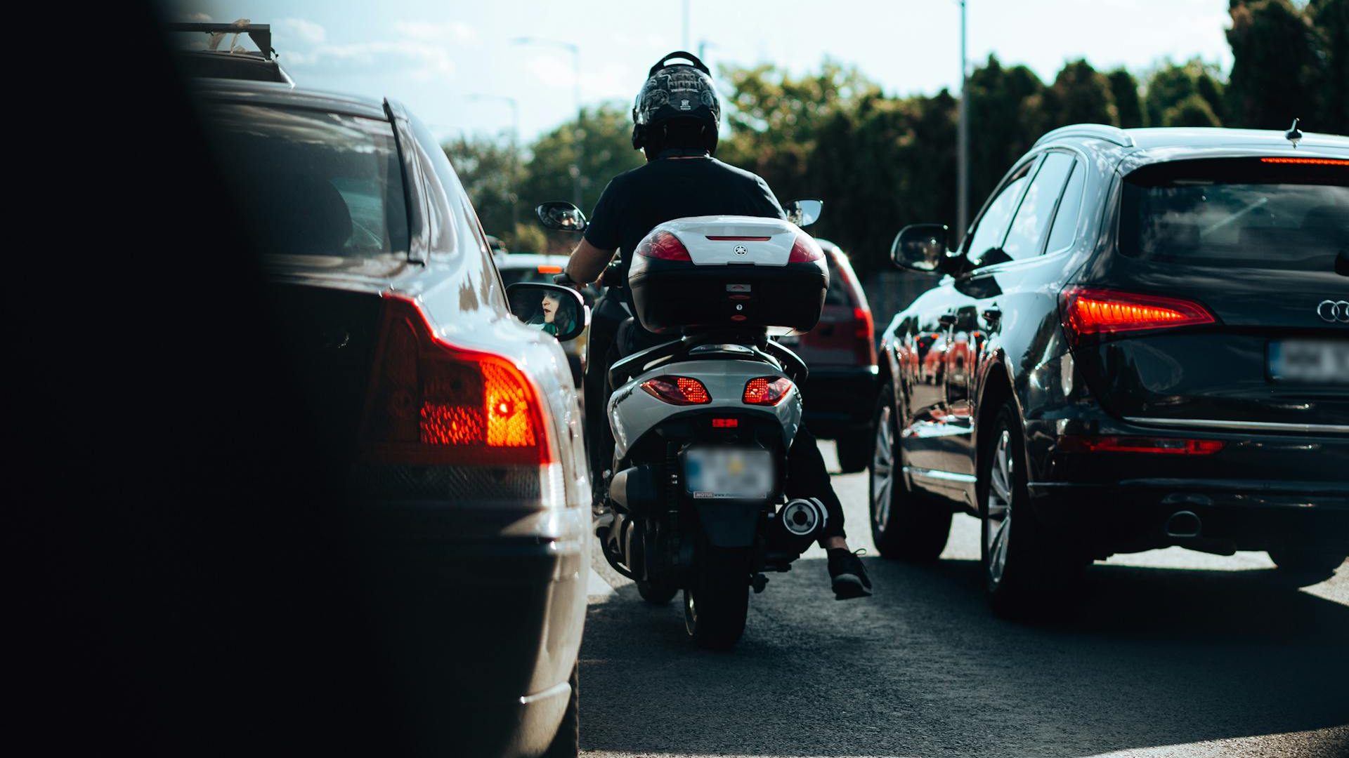 Person on scooter wearing helmet, riding between cars in heavy traffic during daytime.