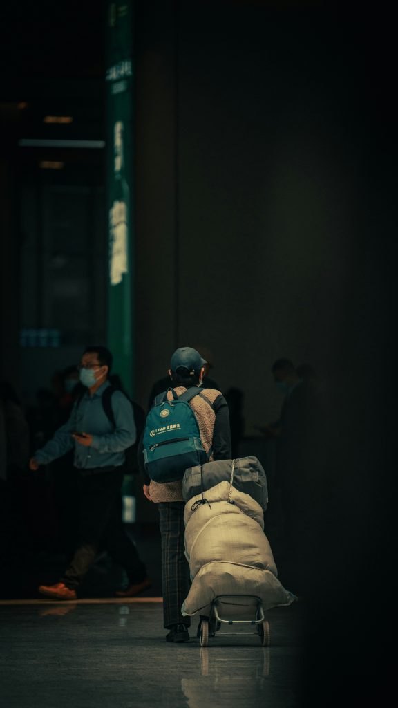 Person with a blue backpack pushes a cart with large bags in a dimly lit indoor public space.