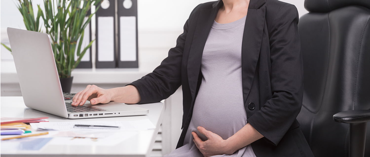 Pregnant woman in business attire working at a desk with a laptop, holding her belly.