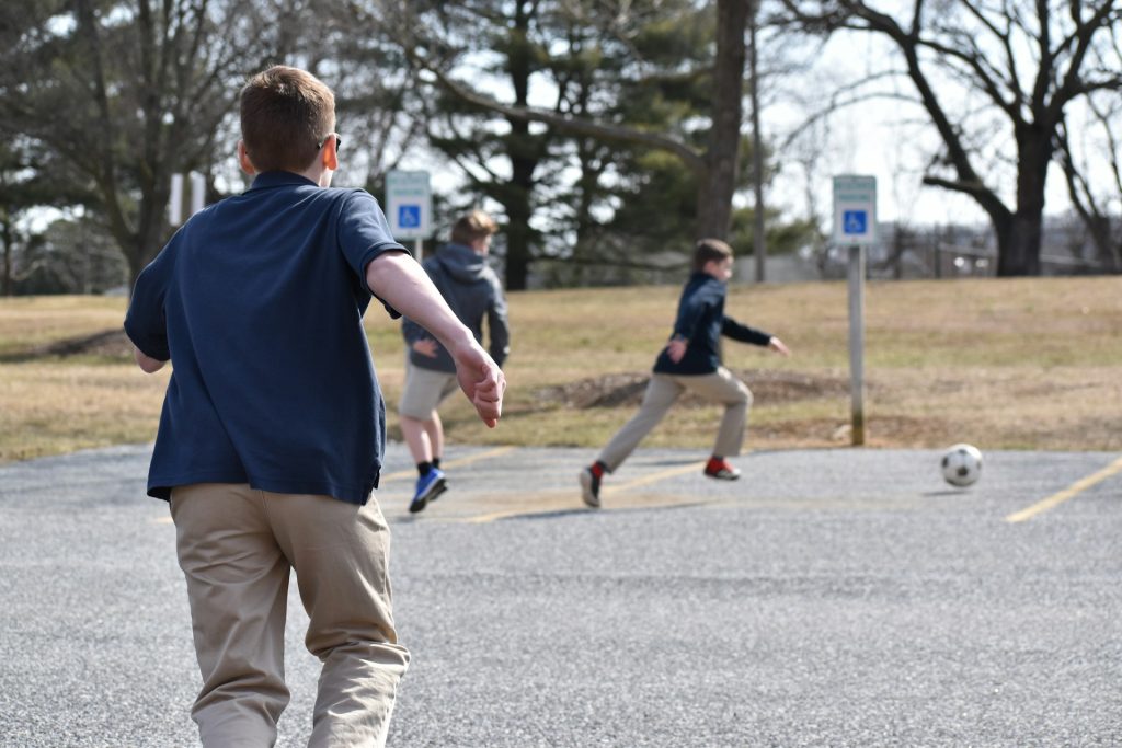 Three boys play soccer in a parking lot on a sunny day, with trees and handicap signs in the background.
