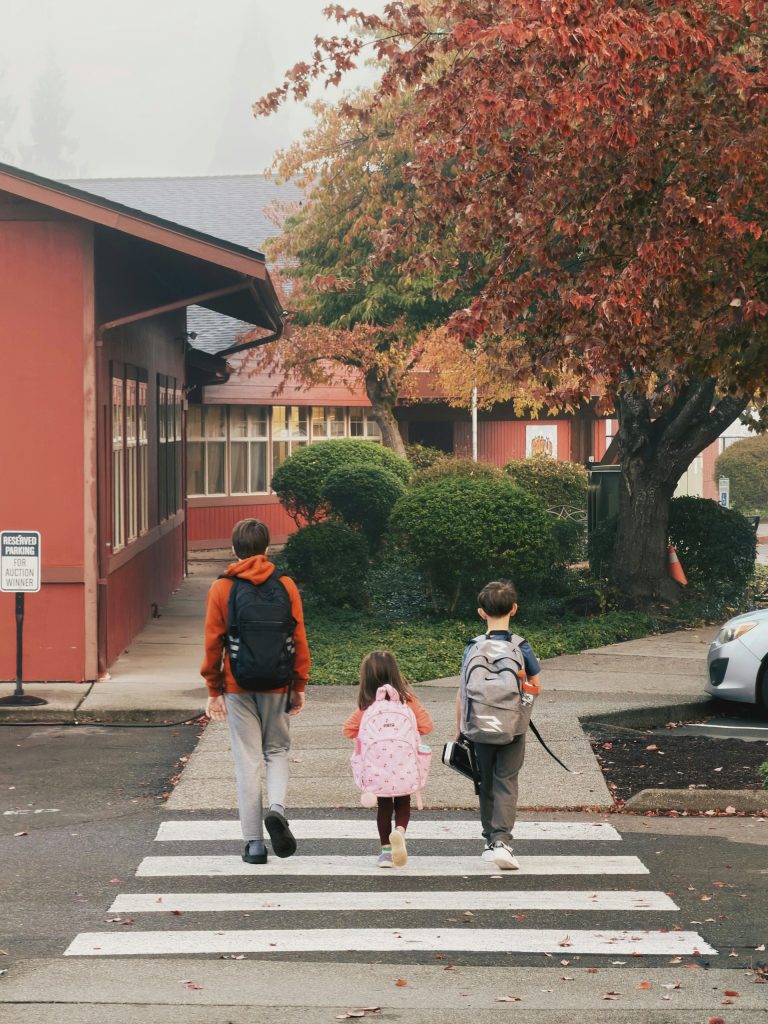 Three children with backpacks cross a street toward a school building lined with autumn trees.