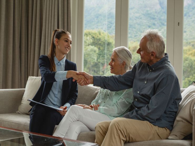 Young woman in a suit shakes hands with an older man while an older woman sits beside him on a couch.