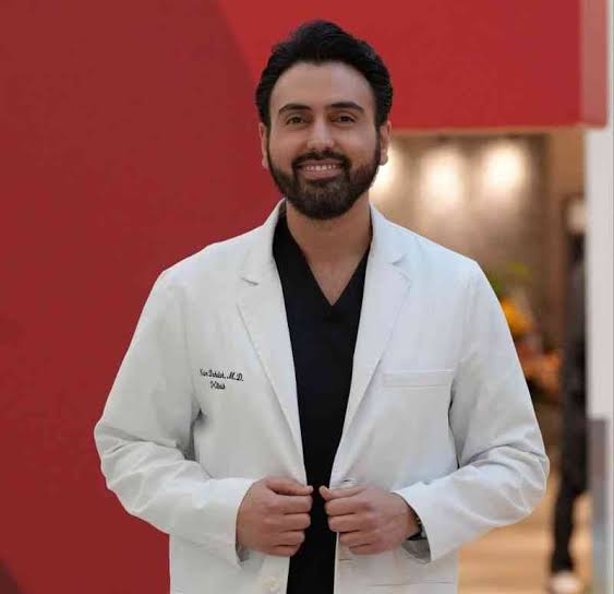 A man in a white lab coat smiles and stands against a red and gray indoor background.