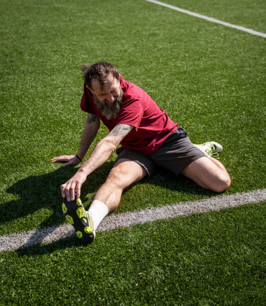Man in red shirt stretching his leg on a grassy sports field near a white line.