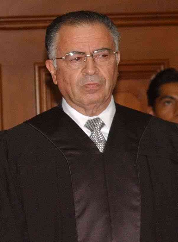 Older man in a black robe and tie stands in a wood-paneled room, looking slightly to the side.