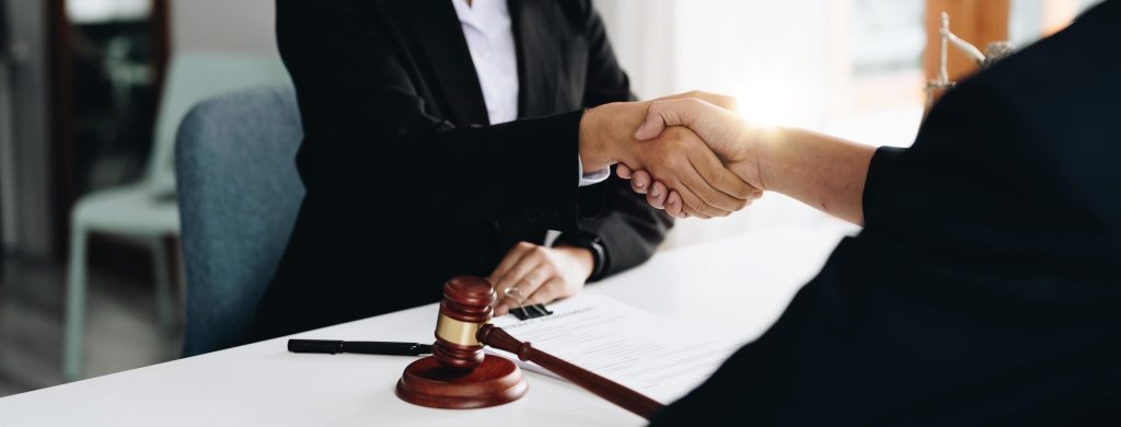 Two people in business attire shaking hands at a desk with a gavel, document, and pen visible.