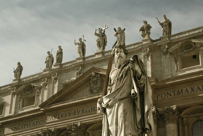 Statue in front of a historic building topped with more statues, with Latin inscriptions on the facade.