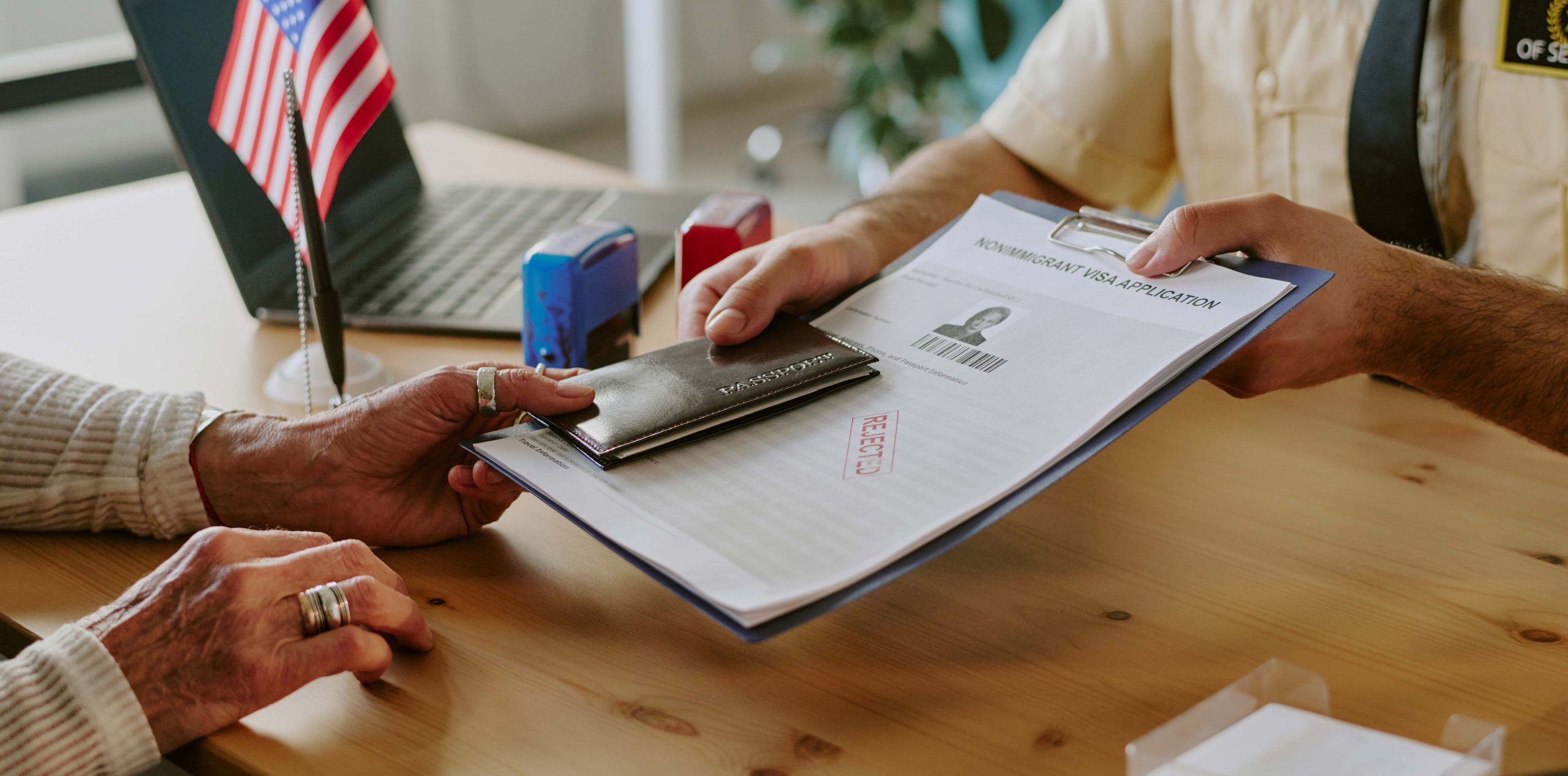 Two people exchange a passport and visa application form at a desk with a laptop and an American flag.