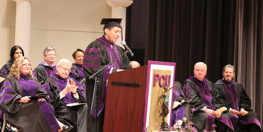 A graduate in academic regalia speaks at a podium labeled “PCU” while faculty members sit behind him.