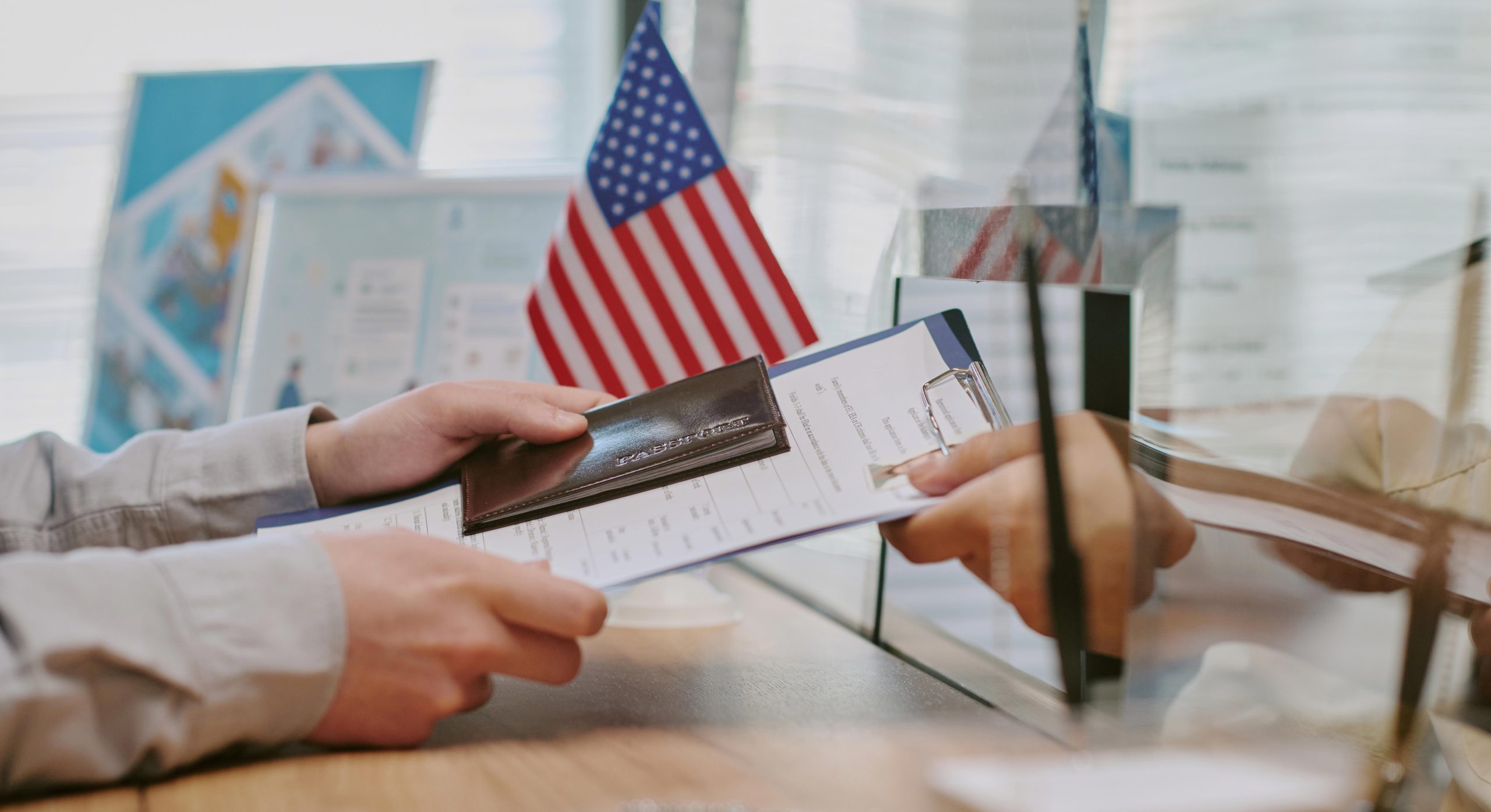 A person hands a passport and a document to a clerk through a glass window.