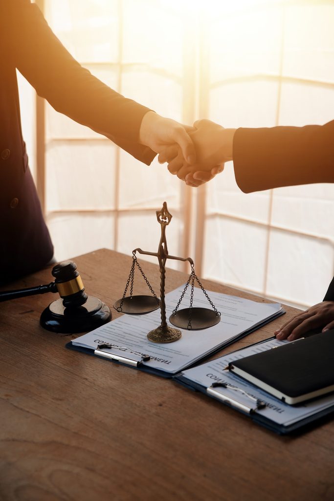 Two people in suits shaking hands over a wooden table with a gavel and a scale of justice.