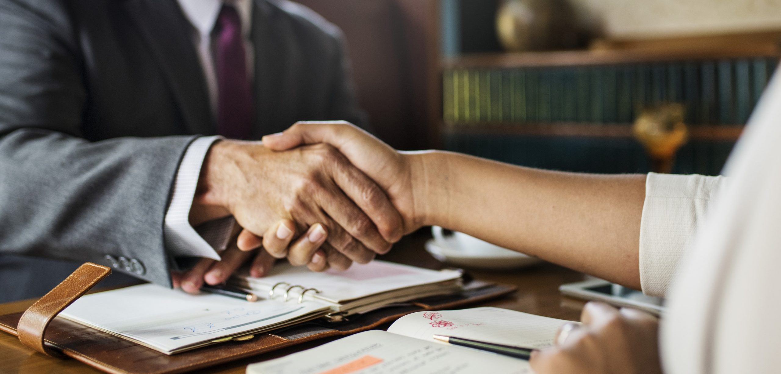 Two business professionals shaking hands over a desk with open notebooks.